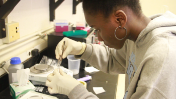 A black, female student in gloves and lab coat looks in a microscope.