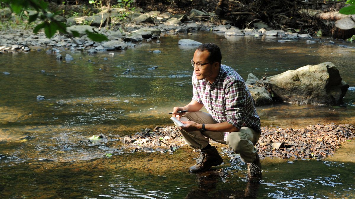 Student crouches at the edge of a river to test water quality