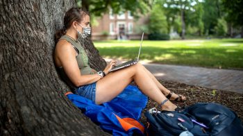 A student works on her laptop while sitting against a tree.