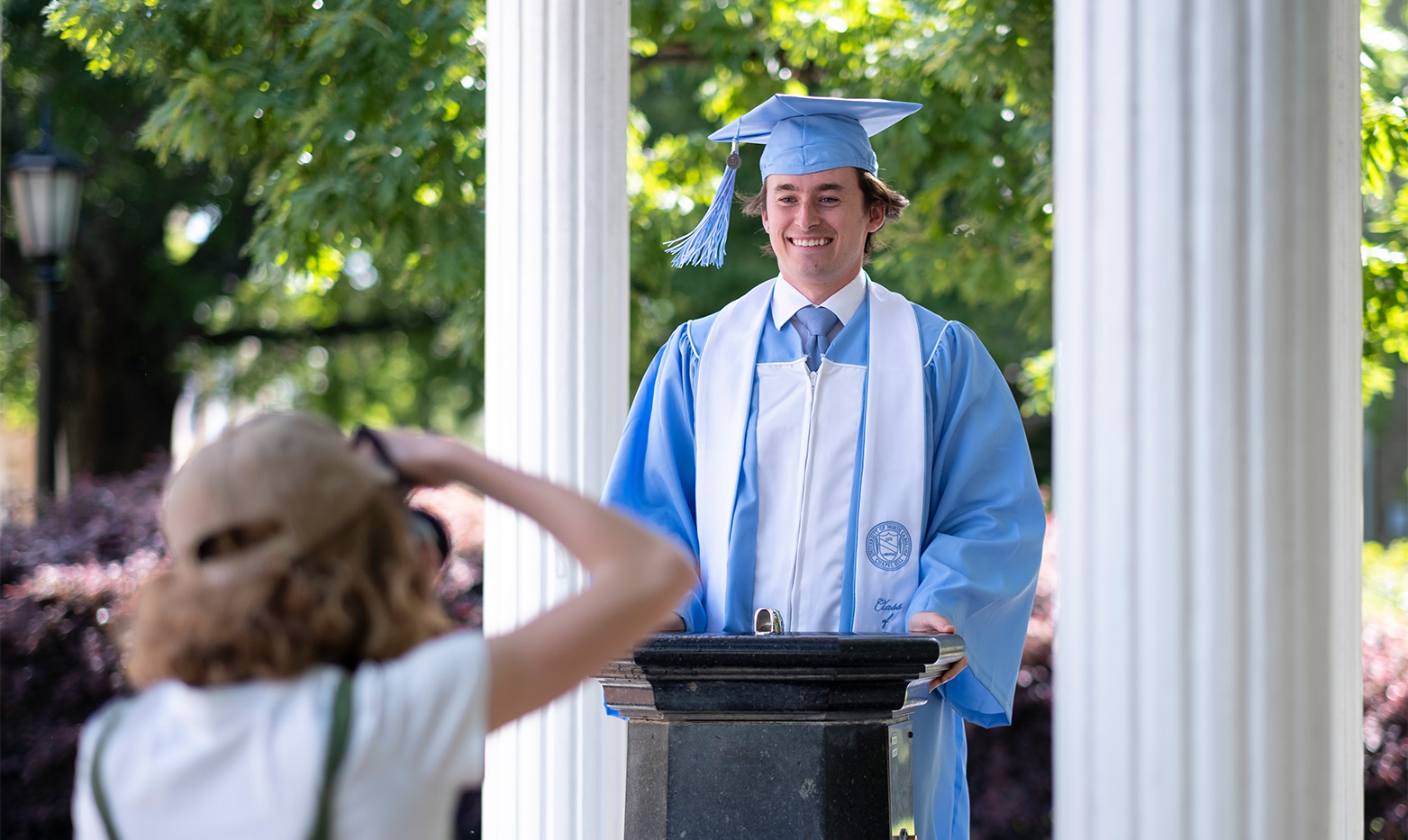 A UNC student in graduation regalia stands at the fountain of the old well.