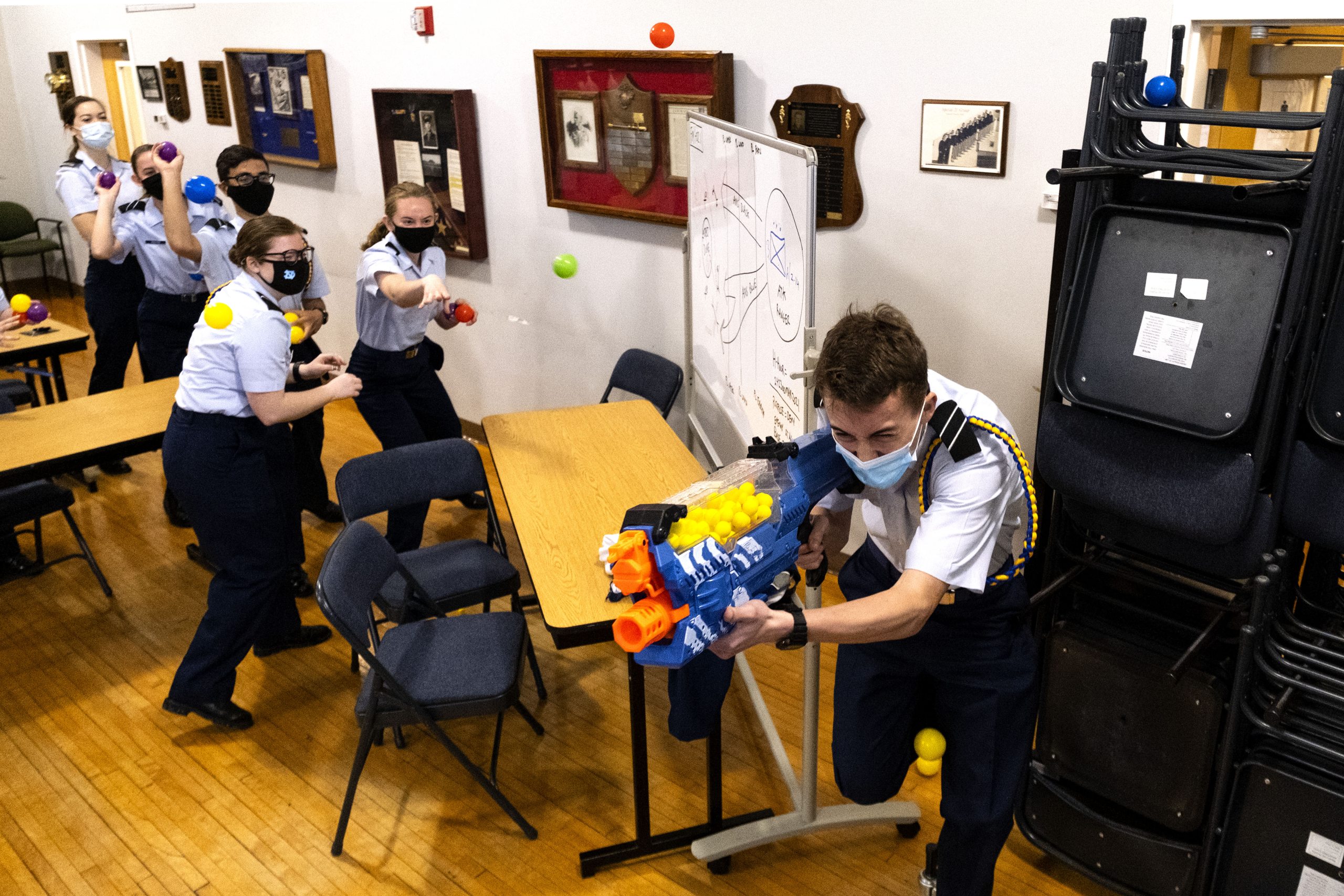 Five students throw balls at Nerf-gun-toting student as he ruuns away. All wear Covid masks.
