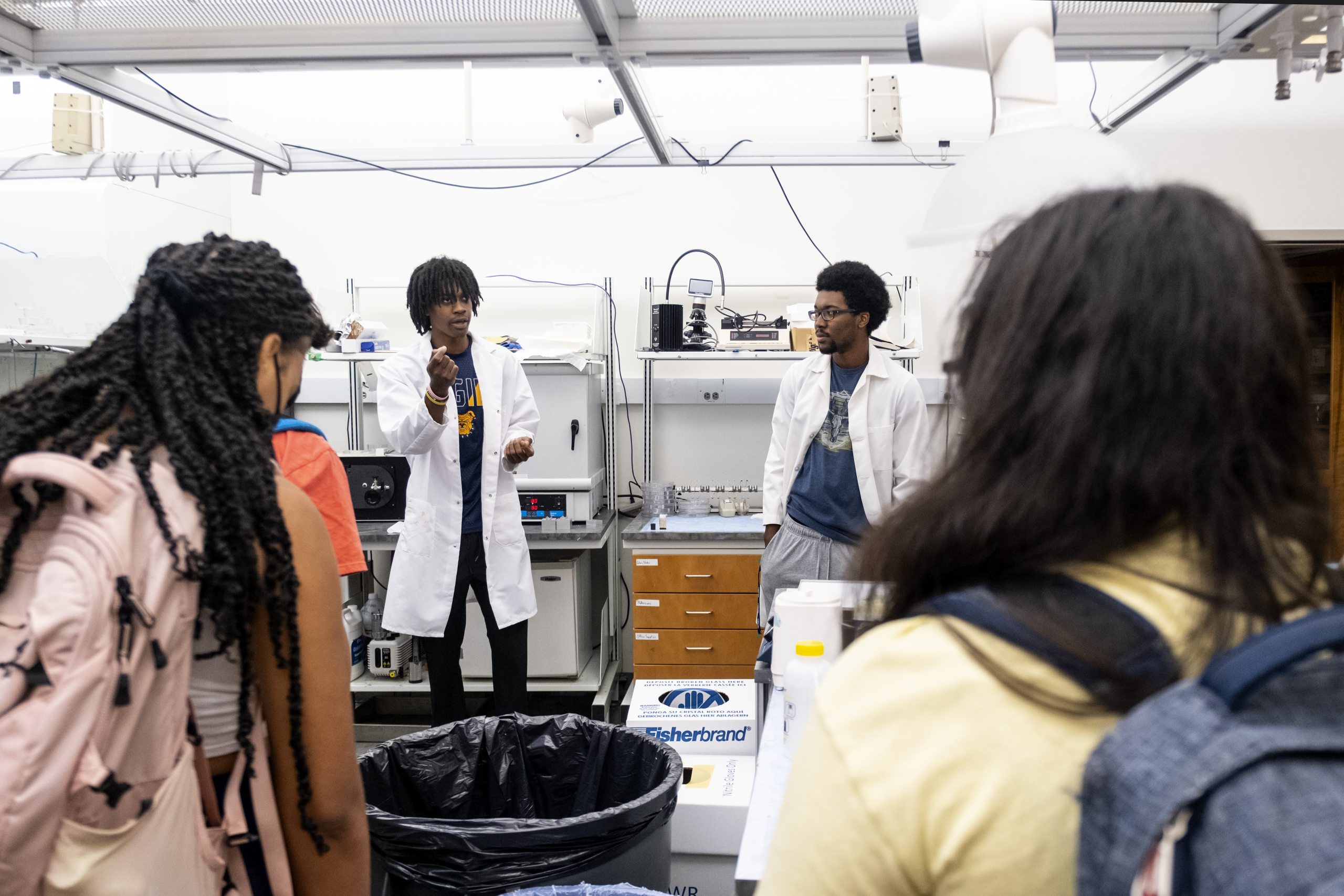 Two Black student researchers in white lab coats explain their research to rising senior high school students.