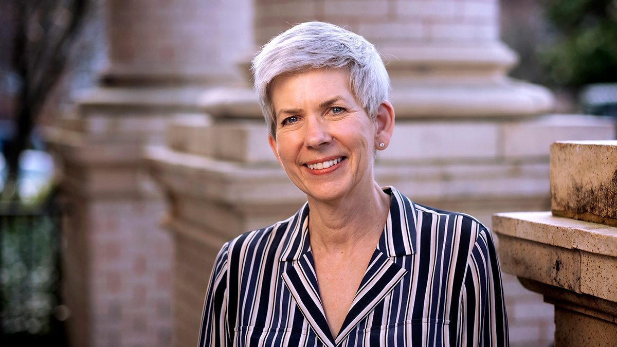 Dean Beth Mayer-Davis smiling at the camera in a striped collared shirt with architectural columns in the background.