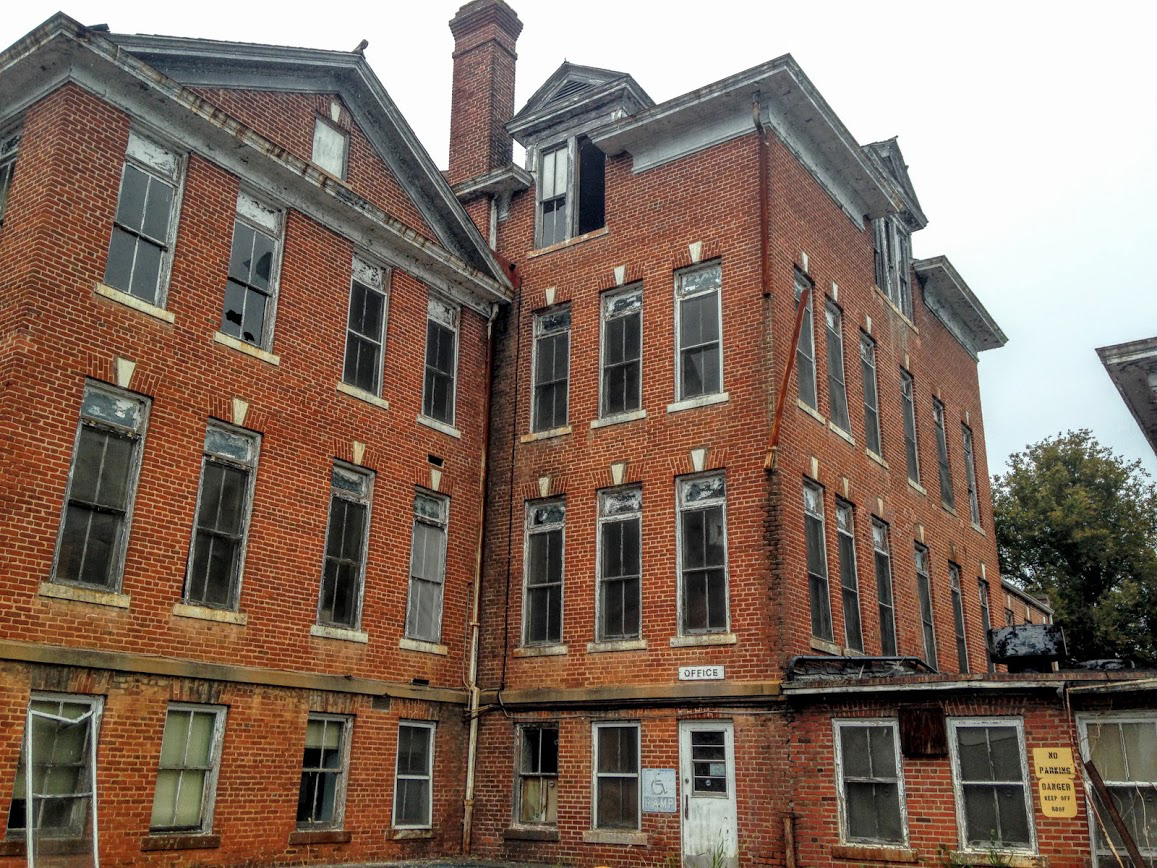 Back side of a Broughton Hospital building showing broken windows and screens and a dingy exterior.
