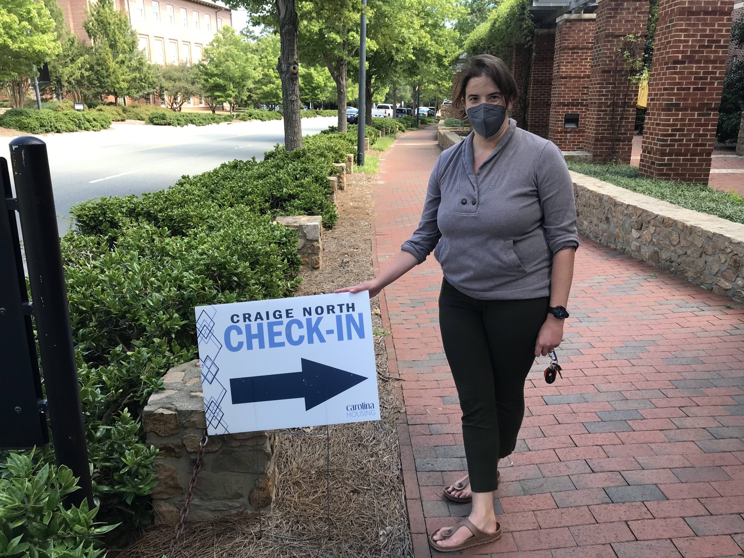 Woman in mask stands beside a sign that reads: Craige North check-in