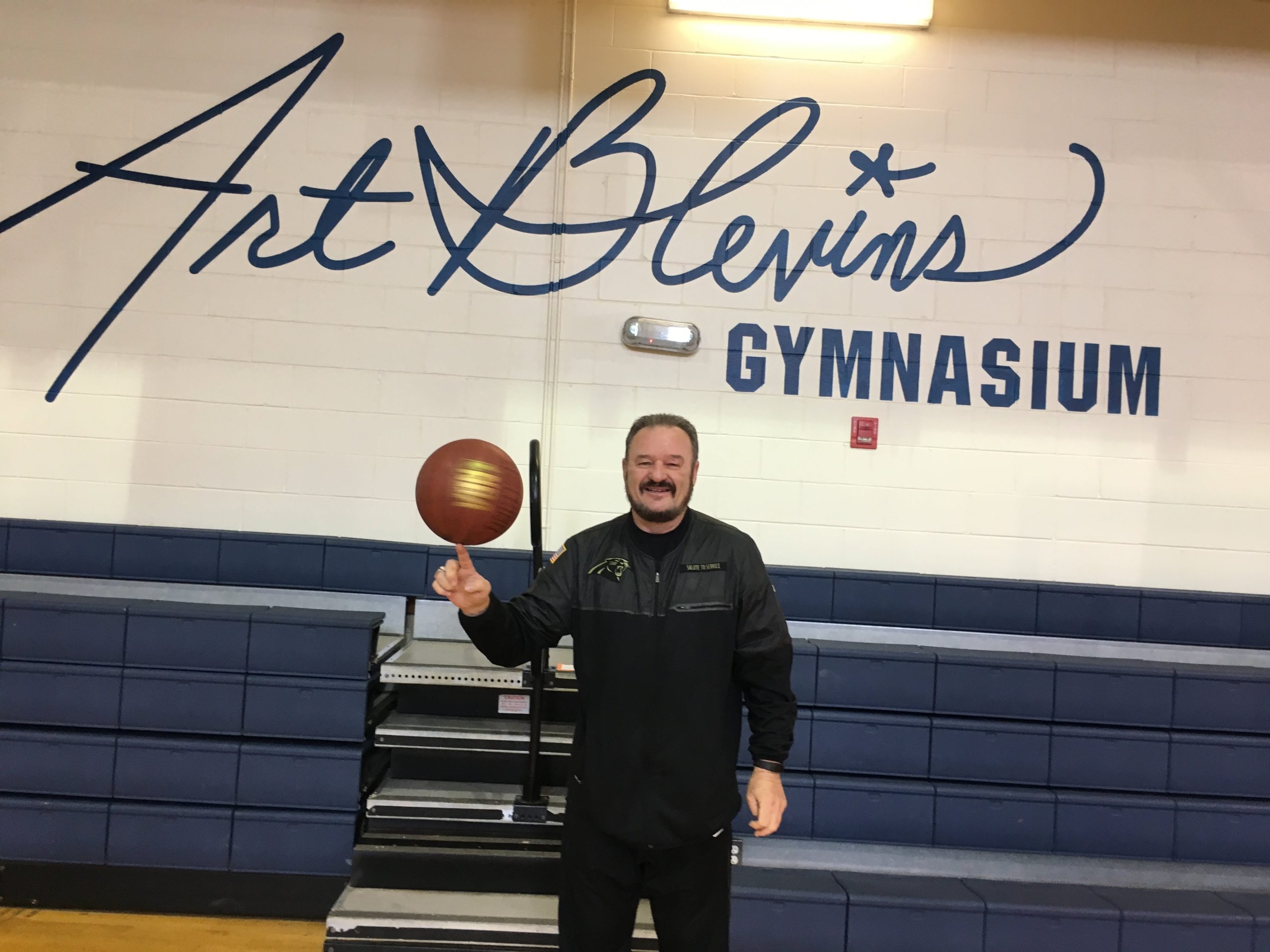 Bearded man spinning a basketball in a gym with "Art Blevins Gymnasium" painted in large letters on the wall behind him.