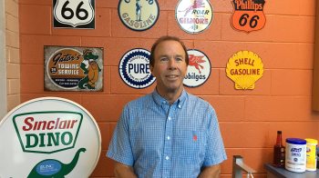 Mark Stark in front of an orange shop wall hung with automotive product signs.
