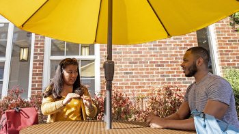 Woman speaking with man at table with a yellow umbrella outside.