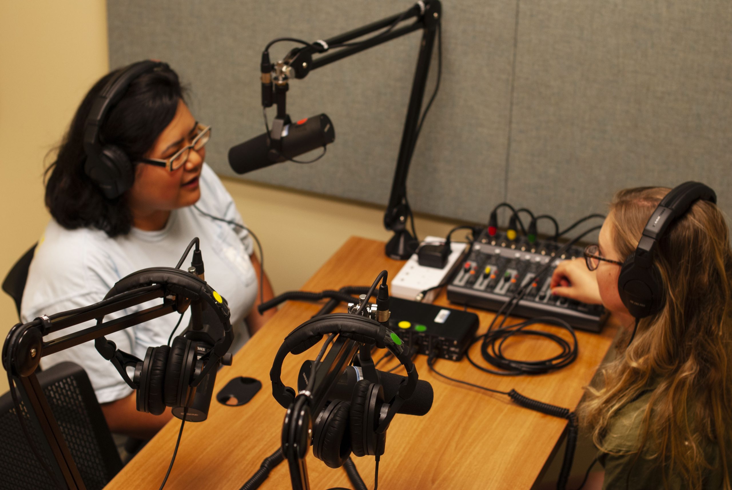 Two women at desk, one speaking into overhead microphone, in podcast studio.