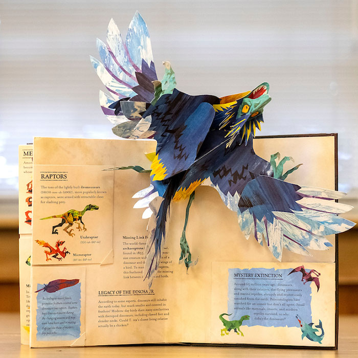 A paper 3D raptor "flies" off a page of a pop-up book.