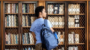 A student standing in a library with his backpack hanging over his right shoulder as he looks to his right in front of a bookcase.
