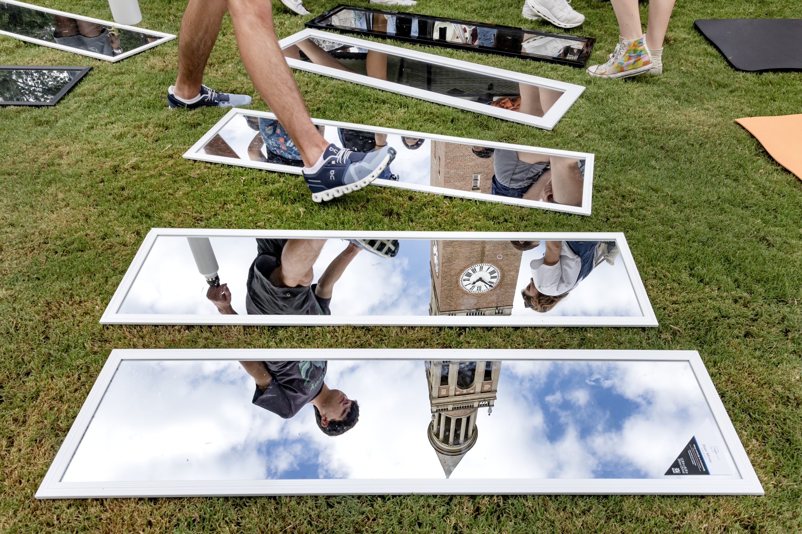 A person stepping around four mirrors sitting on a grass lawn. The reflection of the mirrors creates an upside down view of a bell tower on the campus of UNC-Chapel Hill.