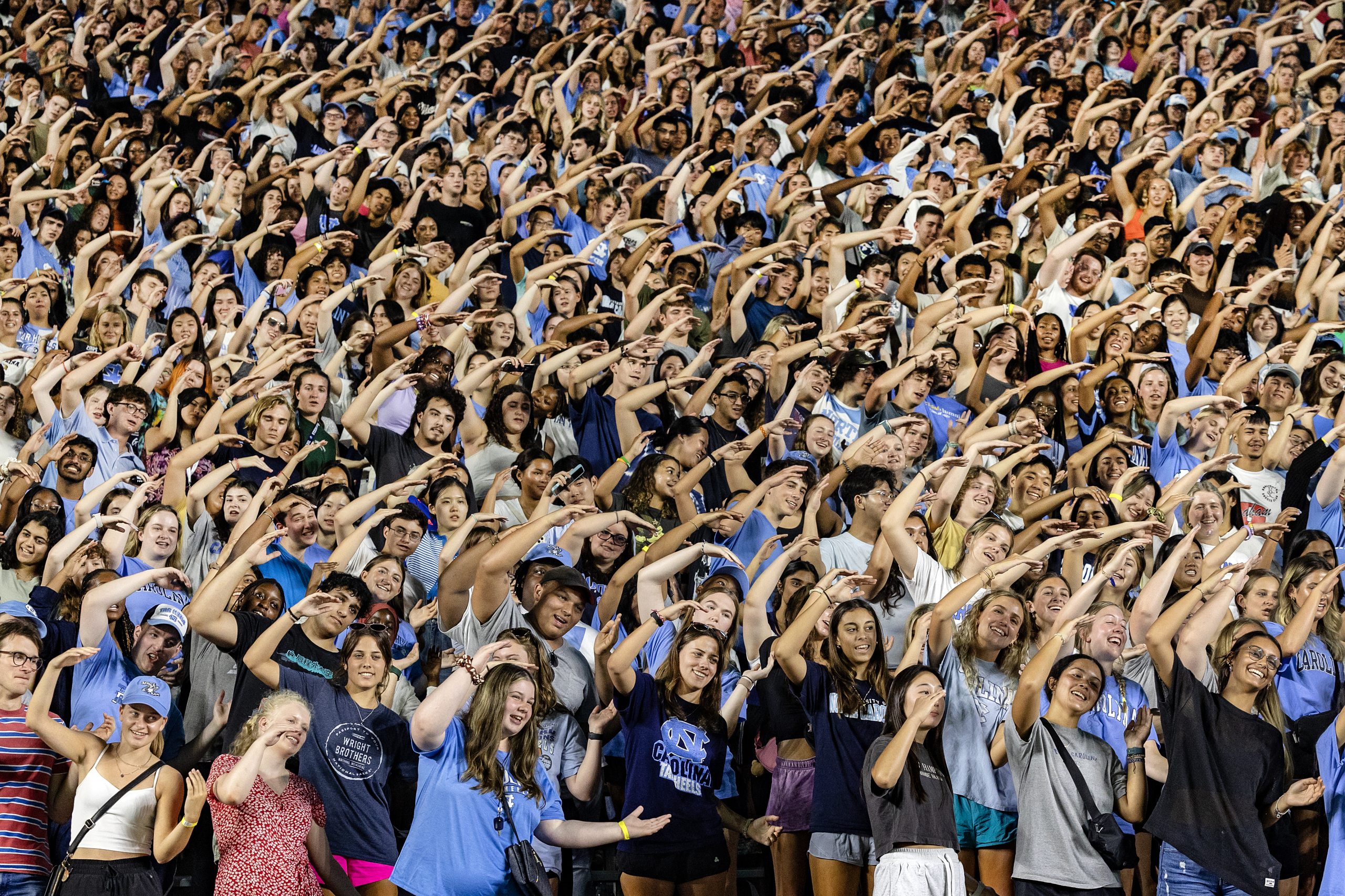 A large crowd of students in a football stadium turning their bodies and positioning their arms to spell the letter 