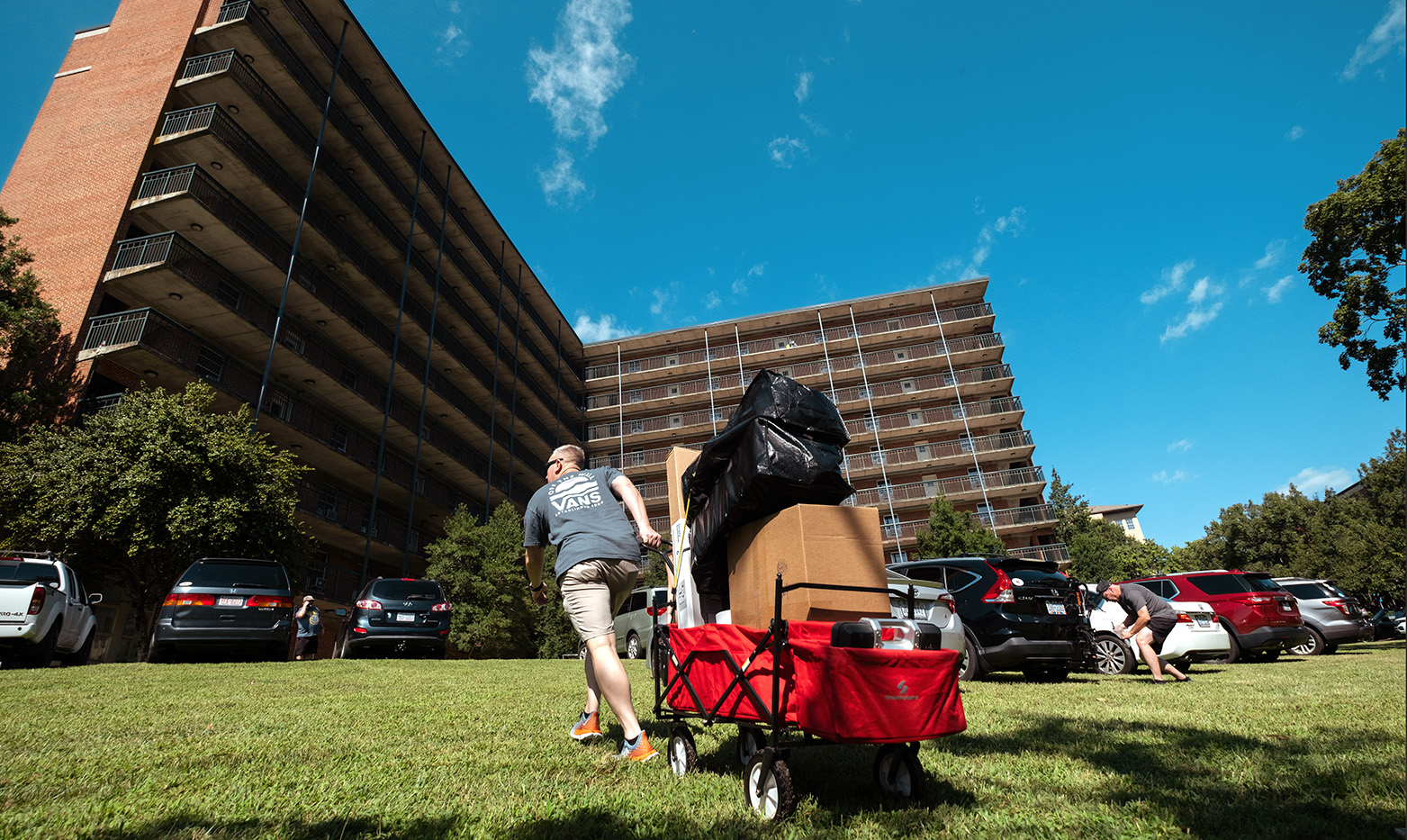 A man using a wagon to transport boxes and bags on a lawn outside of a dormitory.