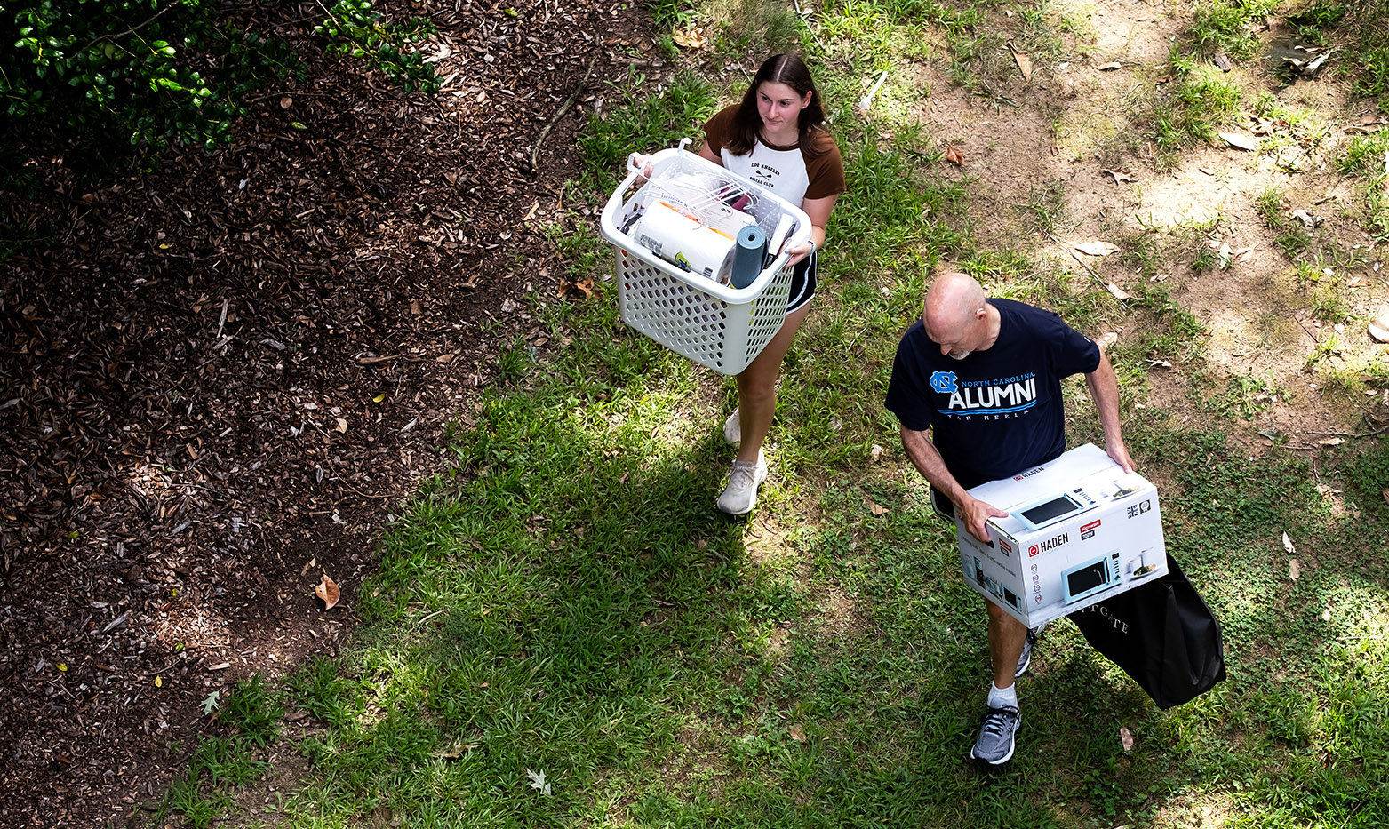 Aerial image of a student and a parent carrying supplies as they walk on the grass outside of a dormitory.