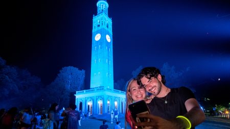 Two students taking a selfie in front of a bell tower lit Carolina Blue at night.