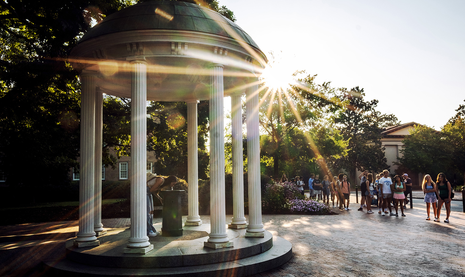 A student drinking from the fountain of the Old Well on the campus of UNC-Chapel Hill. A line of students is seen in the background as the sun shines.