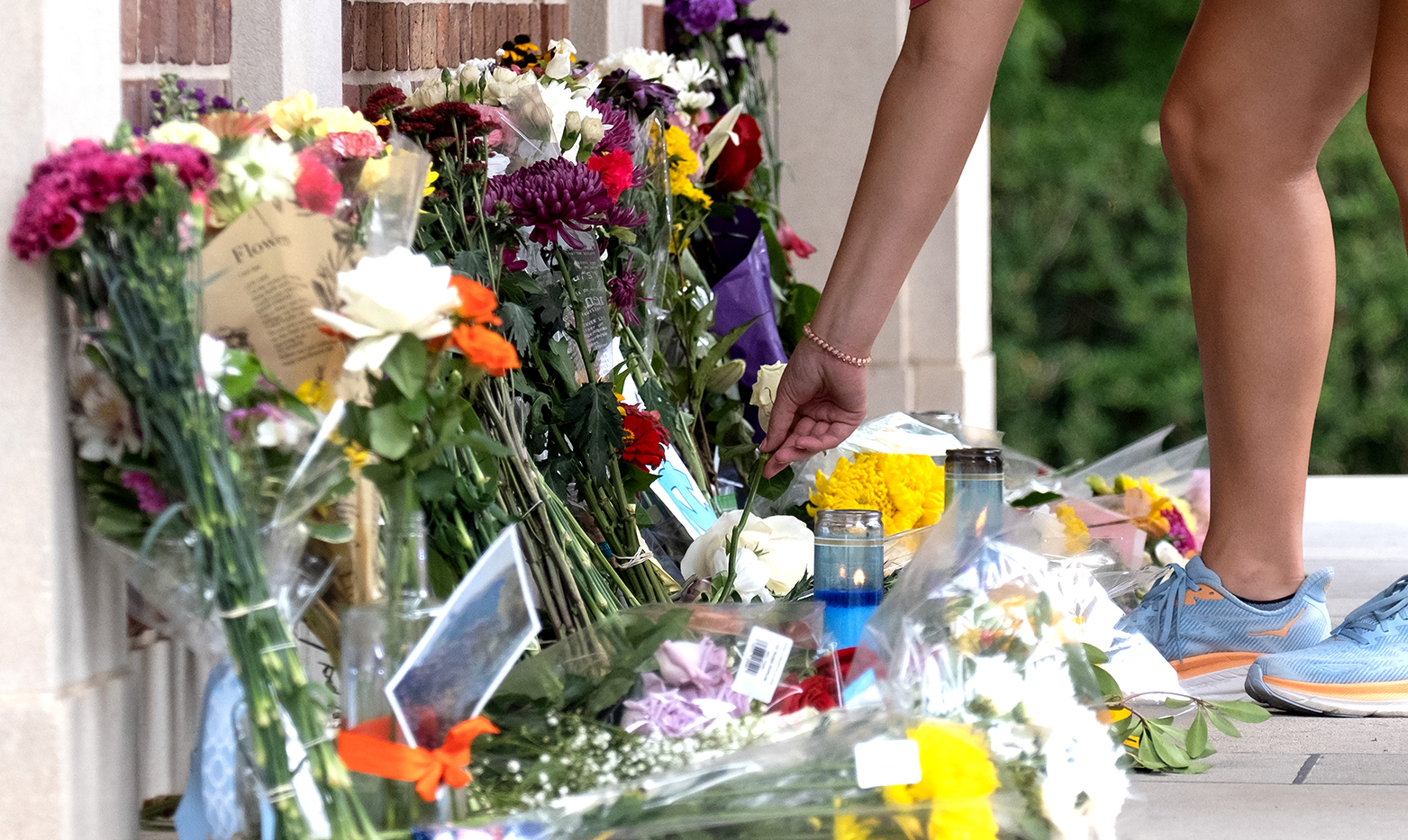 A person's hand placing flowers at a makeshift memorial at the base of a bell tower.