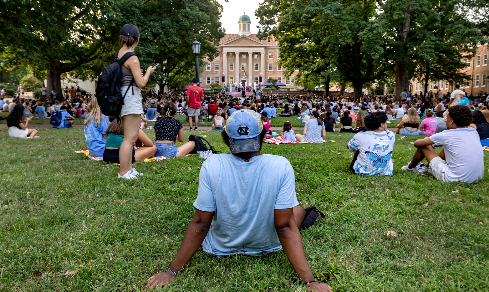 A student, with his back facing the camera, sits on the lawn of Polk Place among a crowd of other students watching a cappella groups perform.
