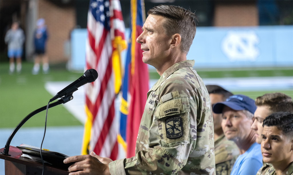 A U.S. Army member speaking into a microphone at a lectern on a football field. Four people sitting behind him to his right focus their attention on him as he speaks.
