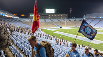 Four people wearing Carolina Blue shirts and backpacks climbing up stadium stairs. Two of them hold flags, one of which reads 