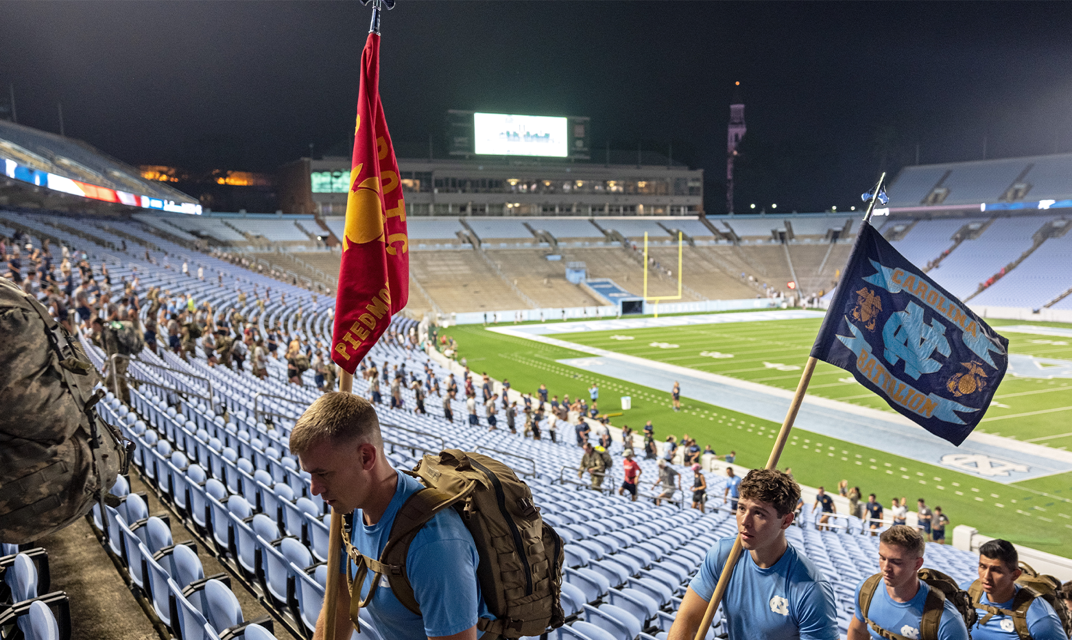 Four people wearing Carolina Blue shirts and backpacks climbing up stadium stairs. Two of them hold flags, one of which reads 