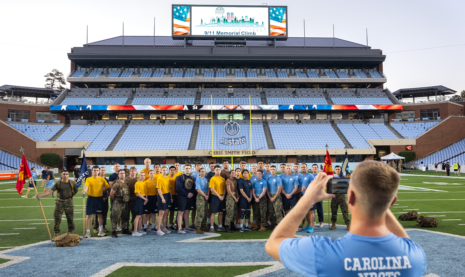 The back of a person holding a photo horizontally and taking a group photo of ROTC members on a football field. In the background, a video board shows a graphic with American flags reading 