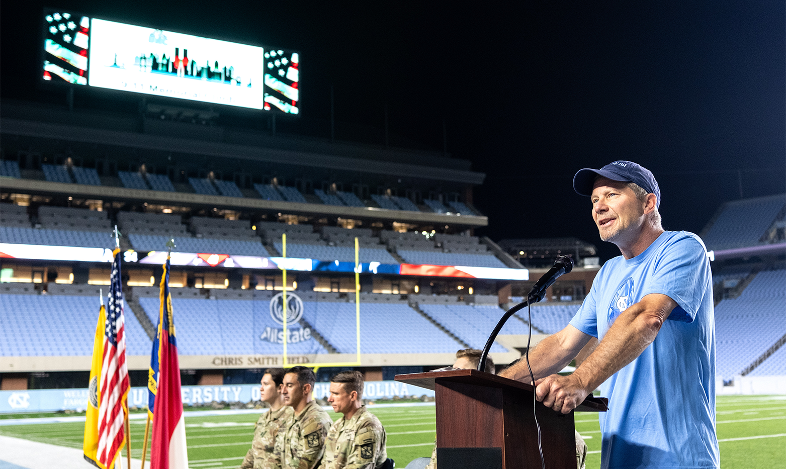 A man standing at a lectern and speaking into a microphone on a football field. Four military members sit slightly behind him to his right, where there are also three flags, including the state of North Carolina flag and the American flag.