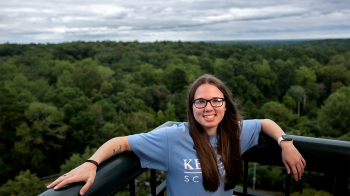 A student, Emily Shipway, posing for a photo on the balcony of a dormitory with a gray sky and an aerial view of green trees in the horizon.