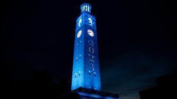 Nighttime photo of the Bell Tower on the campus of UNC-Chapel Hill lit Carolina Blue with projected, white text reading 