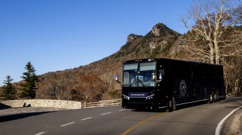 A large navy blue bus driving on a a roadway with a mountain and trees in the background.