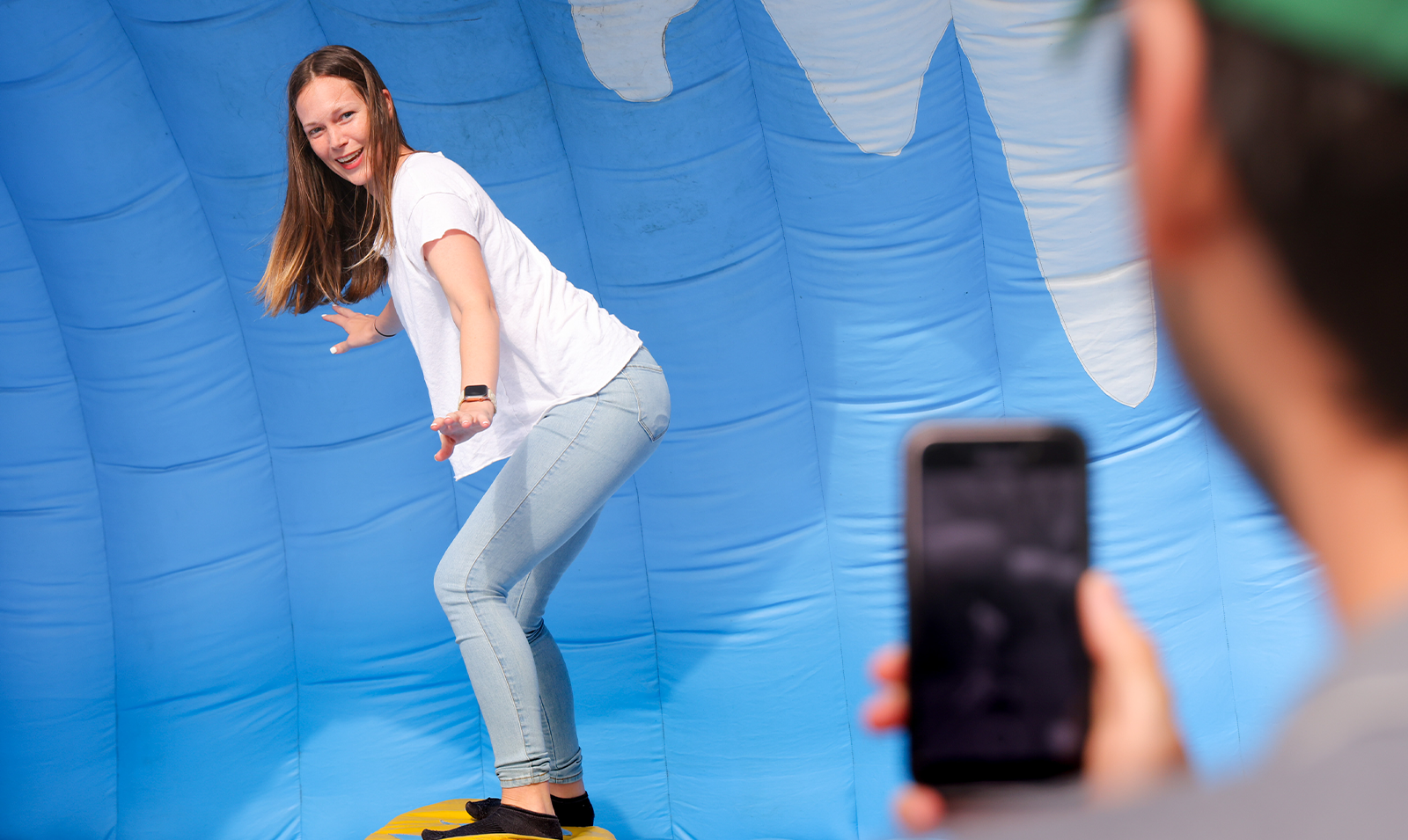 A woman riding on a surf simulator while a person in the foreground takes a picture on an iPhone.
