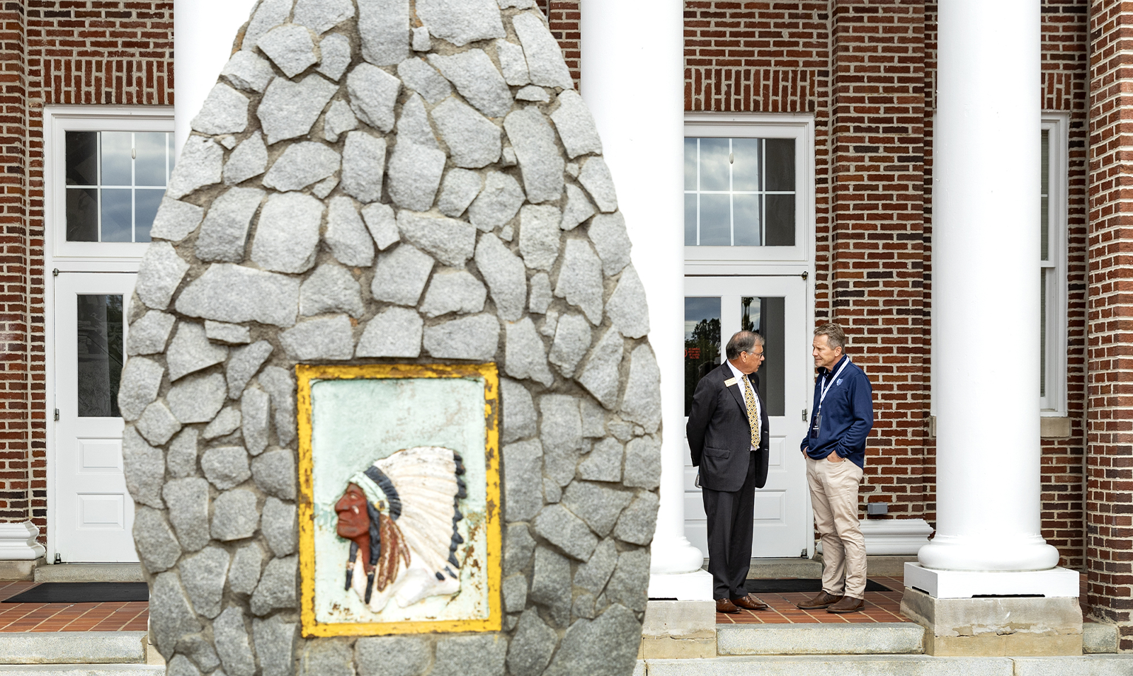 Two men chatting outside of a building in the background. In the foreground is a painting of an American Indian atop a stone sculpture.