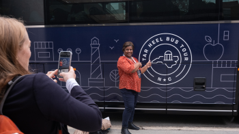 A person using an iPhone to take a picture of a woman posing next to a large bus and pointing to the 