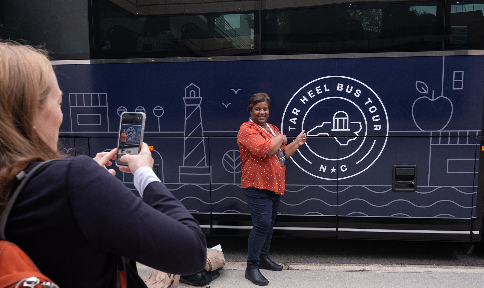 A person using an iPhone to take a picture of a woman posing next to a large bus and pointing to the 