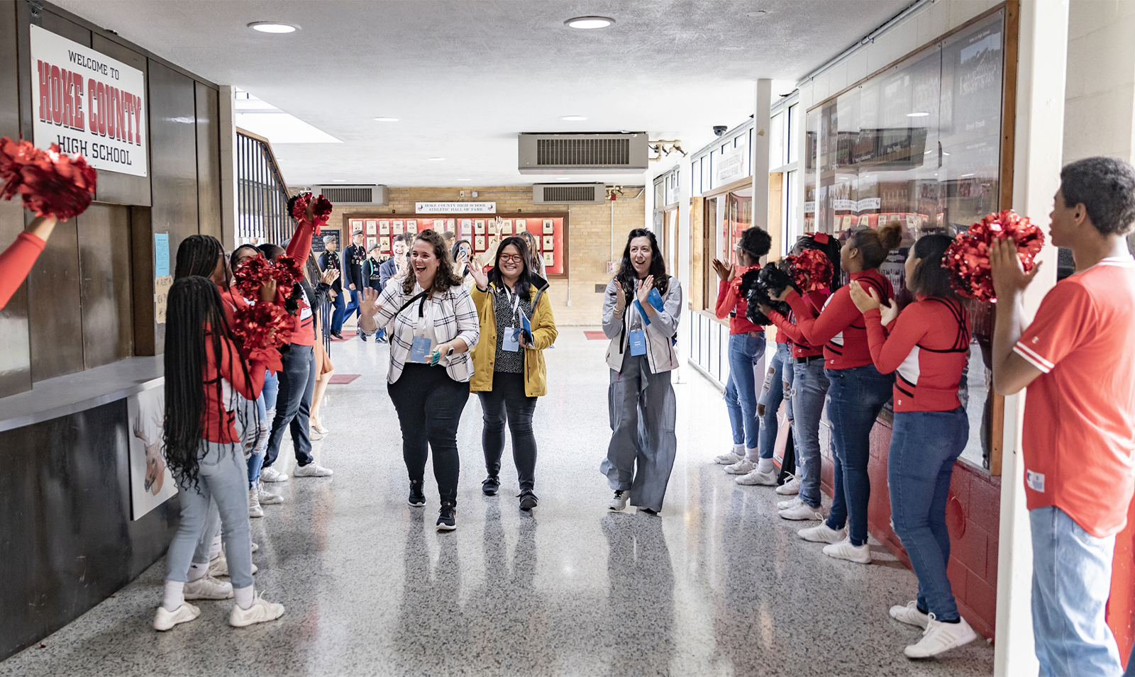 Cheerleaders with pom-poms cheering as school guests walk through a hallway in a high school.