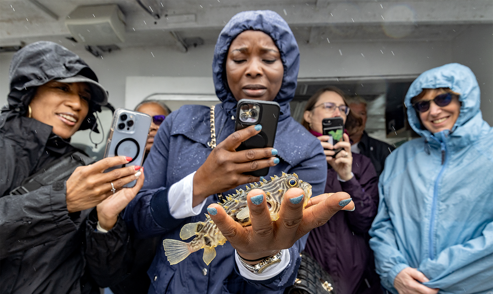A woman on a boat taking a picture with her iPhone of her hand, which is holding a fish.