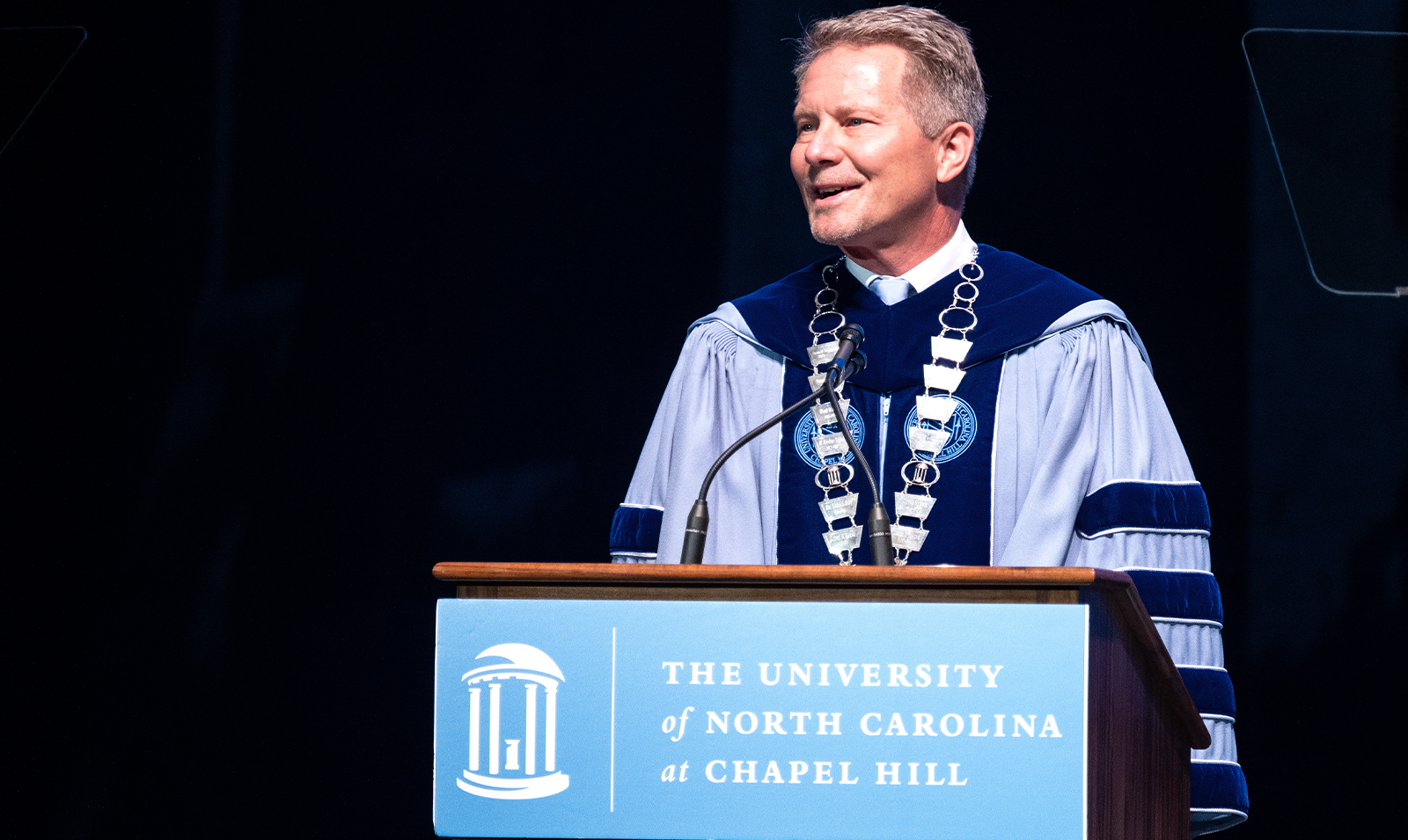 A man, UNC-Chapel Hill Chancellor Kevin M. Guskiewicz, dressed in regalia and speaking at a lectern during a University Day event in an indoor hall.