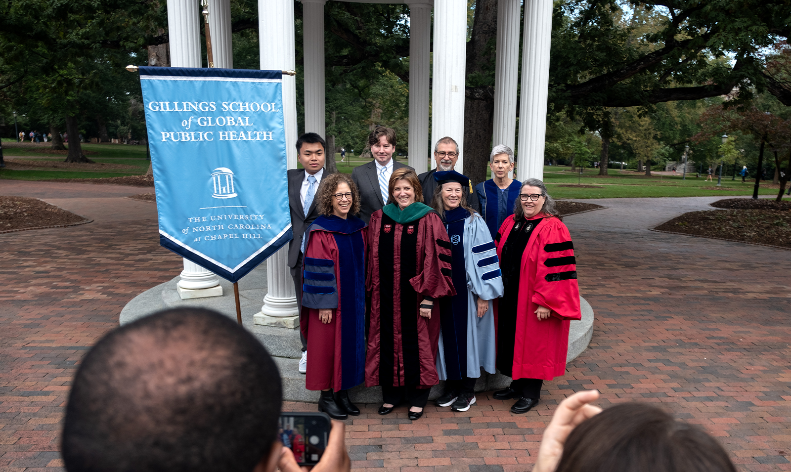A group of peopel, some of whom are in regalia, posing for a group photo in front of the Old Well with a banner that reads 