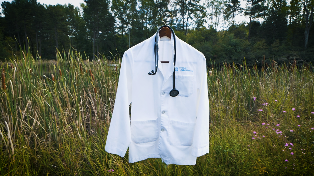 A white coat and stethoscope in front of a rural backdrop, featuring high grass and trees in the horizon.