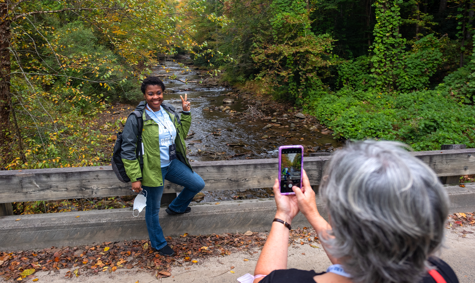 A woman posing for a photo with a peace sign in front of a forested waterway, as another woman takes her picture with an iPhone.