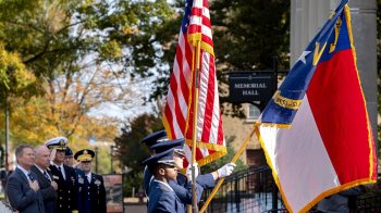 Uniformed ROTC members holding North Carolina and American flags as part of a Veterans Day ceremony at UNC-Chapel Hill outside of Memorial Hall. In the background are men holding their hands across their chest and others in military uniform saluting.