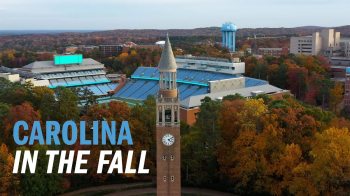 A still image from drone footage of the campus of UNC-Chapel Hill on a fall day. Prominently displayed are the Morehead-Patterson Bell Tower and a Kenan Stadium (a football stadium). In the foreground are trees with orange, brown, yellow and green leaves and a Carolina Blue water tower. Text is overlayed, reading, 