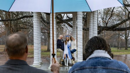 A graduate posing for a photo with family members at the Old Well on the campus of UNC-Chapel Hill on a rainy Sunday, the day of Winter Commencement. In the foreground is a man holding a Carolina Blue and white umbrella.