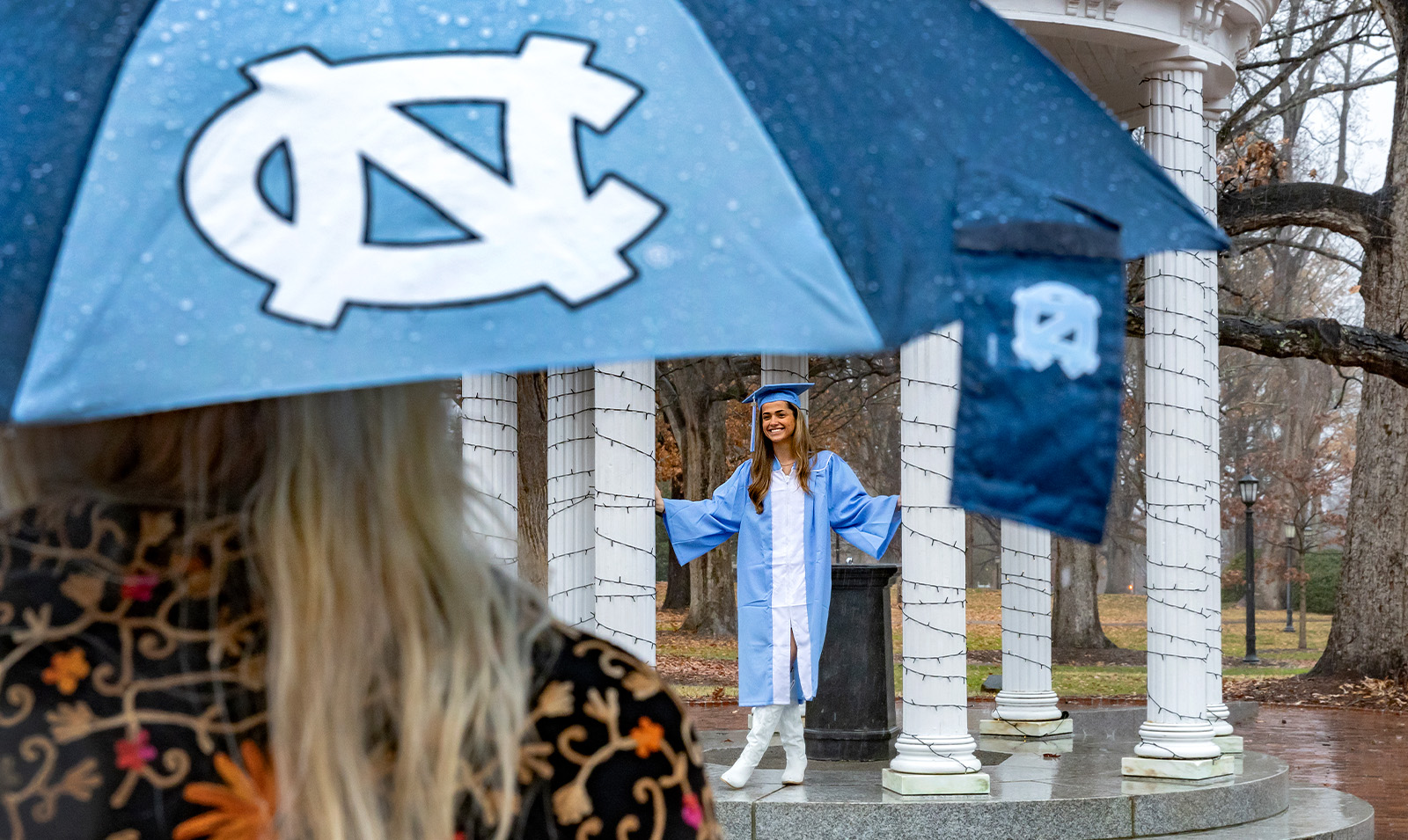 A graduate posing for a photo at the Old Well on the campus of UNC-Chapel Hill on a rainy Sunday, the day of Winter Commencement. In the foreground is a man holding a Carolina Blue and white umbrella.