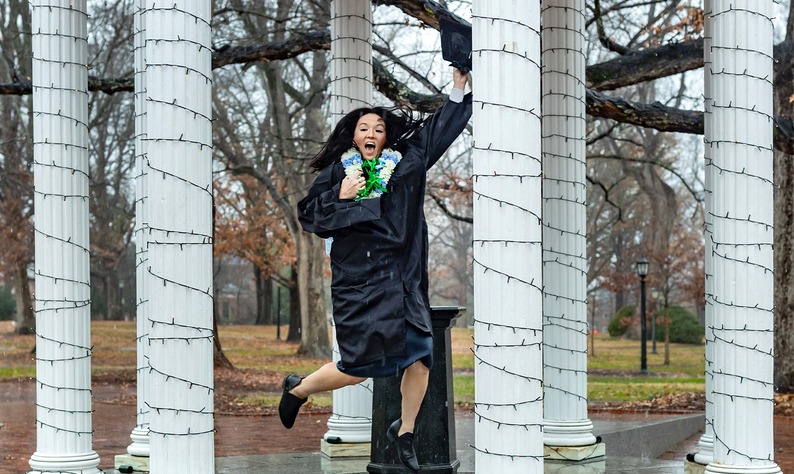 A graduate jumping up in the air and smiling in front of the Old Well.