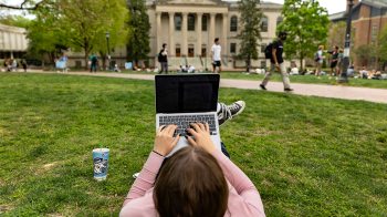 Student laying down on the main quad with a laptop placed on their legs.