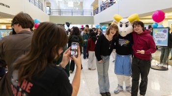 A person taking a picture of two people posing with a ram mascot, Rameses.
