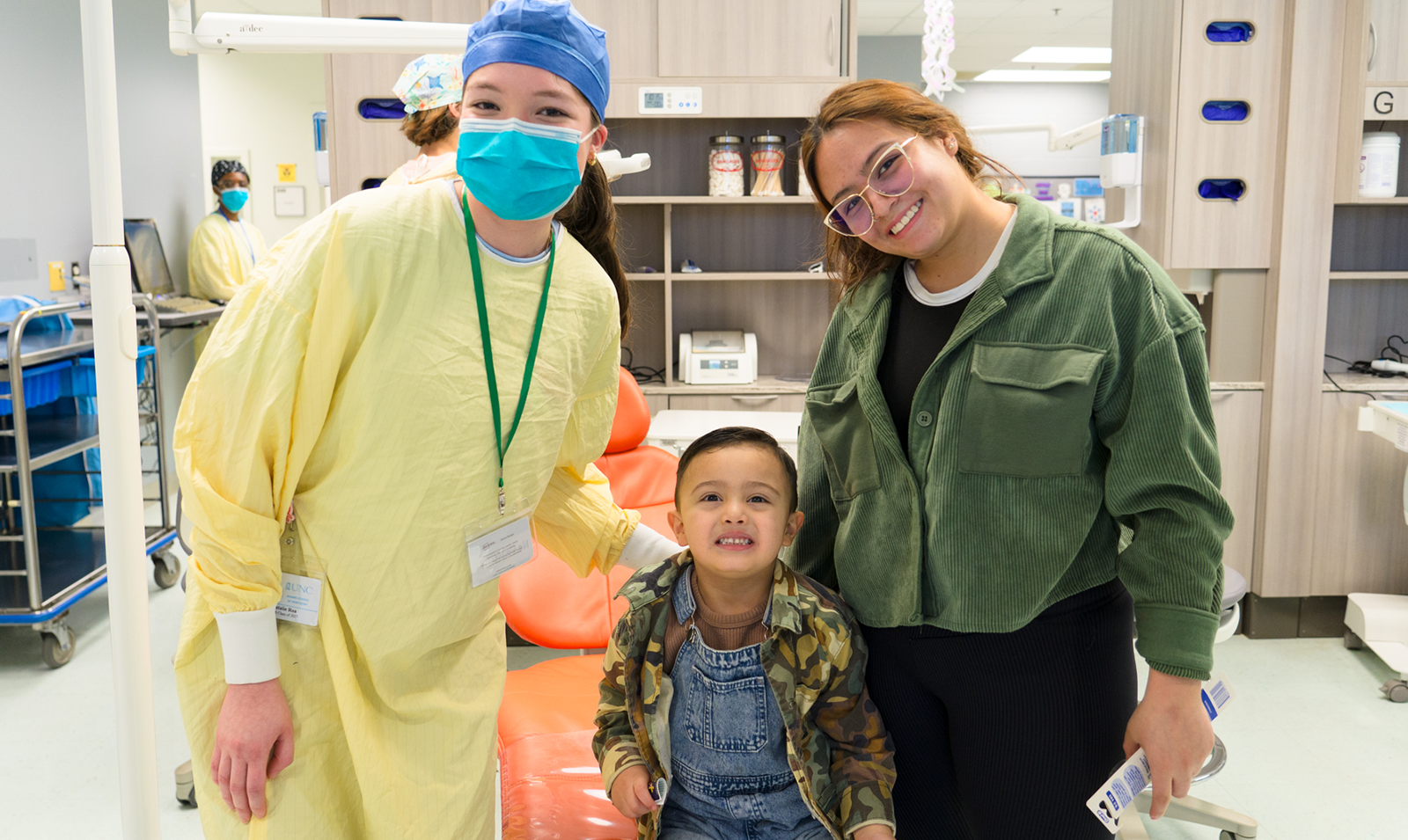 Nurse in scrubs poses with woman in green jacket and smiling child in the middle.