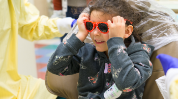 Child with curly hair wearing sunglasses and a sweatshirt with butterfly designs all over it.